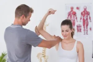 Chiropractor examining patient's arm and elbow range of motion in clinical setting with anatomical charts on wall