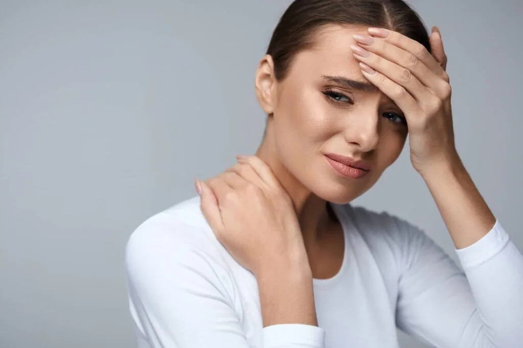 Woman in white long-sleeved shirt touching her forehead with one hand and her neck with the other, appearing to experience discomfort or pain
