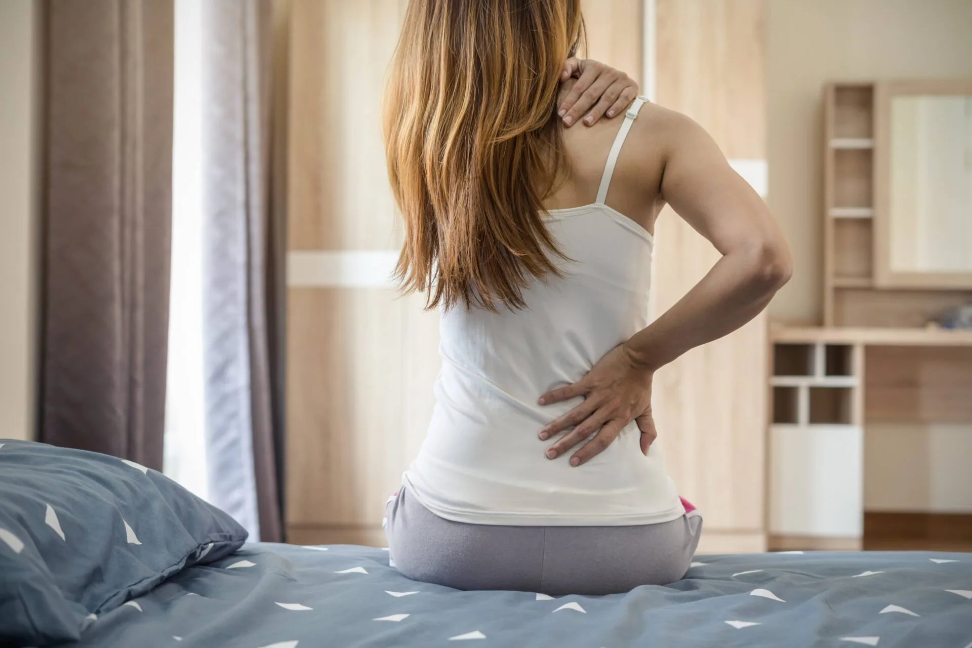Woman sitting on bed touching her lower back and neck in discomfort